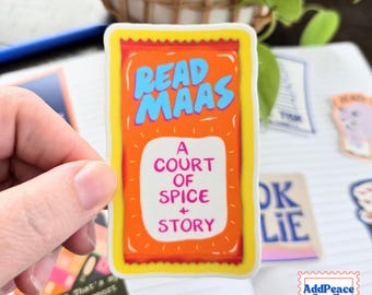May include: A rectangular sticker with a bright orange background and a yellow border. The text READ MAAS is printed in blue at the top, and the text A COURT OF SPICE + STORY is printed in pink within a white central box on the sticker.