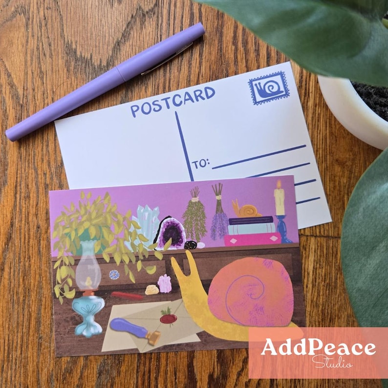 May include: A postcard and an art print with a snail illustration, crystals, and a vintage lamp. The postcard is white with the word "POSTCARD" printed on it. A purple pen rests on the postcard. The art print has the text "AddPeace Studio".