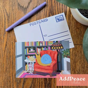 May include: A postcard featuring a cartoon snail in a red armchair, a bookshelf, and a lamp. A blank postcard with the word "POSTCARD" and a pen are on top. The postcard is from AddPeace Studio.