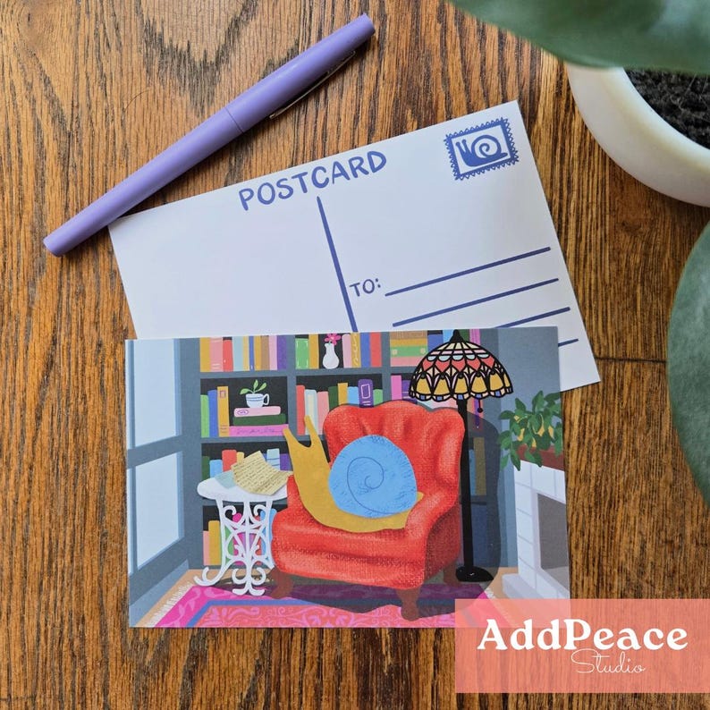 May include: A postcard featuring a cartoon snail in a red armchair, a bookshelf, and a lamp. A blank postcard with the word "POSTCARD" and a pen are on top. The postcard is from AddPeace Studio.