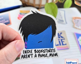 May include: A die-cut sticker featuring a stylized blue book character with messy black hair and a simple line mouth. The text below the character reads, "INDIE BOOKSTORES AREN'T A PHASE, MOM" in a bold, black, hand-lettered font style.