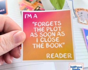 May include: A square sticker with a pink to orange gradient background featuring white text that reads: I'M A "FORGETS THE PLOT AS SOON AS I CLOSE THE BOOK" READER. The sticker has a white border and is displayed against a lined notebook page.