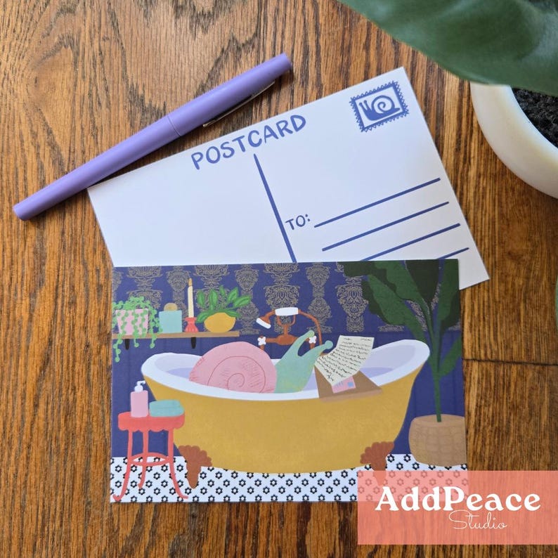 May include: A postcard featuring a cartoon snail in a yellow bathtub, reading. A purple pen and a blank postcard are on a wooden surface. The postcard has the word "POSTCARD" printed on it. The illustration has a blue background.