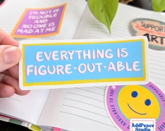 May include: A rectangular sticker with a light blue background, a yellow border, and white text that reads EVERYTHING IS FIGURE-OUT-ABLE. The sticker is positioned over an open notebook with lined pages and other colorful stickers.