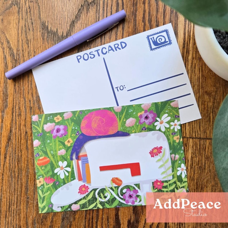 May include: A postcard featuring a snail illustration on a white mailbox amidst flowers. A purple pen and a blank postcard with "POSTCARD" are on a wooden surface. The postcard has the text "AddPeace Studio".