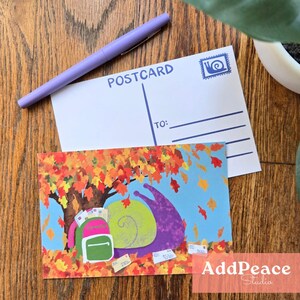 May include: Two postcards and a purple pen are on a wooden surface. One postcard is blank with "POSTCARD" printed at the top. The other postcard features a colourful illustration of a snail with a backpack under a tree with autumn leaves.