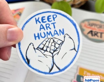 May include: A circular white sticker with a blue border featuring a black ink illustration of two cupped hands. The text "KEEP ART HUMAN" is printed in blue capital letters above the hands. The design is centered on the sticker surface.