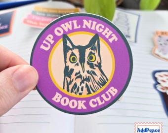 May include: A circular sticker with a purple border and a light peach center featuring a black and white illustration of an owl with yellow eyes. The text UP OWL NIGHT BOOK CLUB is printed in white capital letters around the circular border.