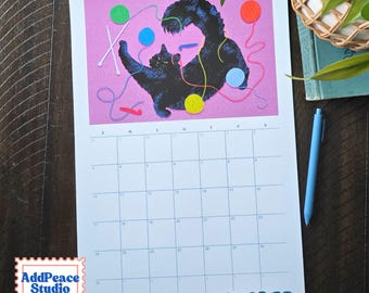 May include: A wall calendar for October featuring a black cat playing with colorful yarn balls and knitting needles on a pink background. The calendar grid is white with black lines and text. A small logo for AddPeace Studio is in the corner.