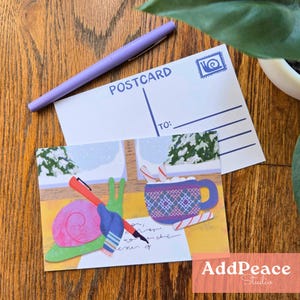 May include: A postcard with a winter scene illustration featuring a snail holding a pen, a mug of hot chocolate, and candy canes. A purple pen rests on top of the postcard. The word "POSTCARD" is printed on the back.