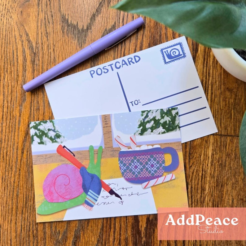 May include: A postcard with a winter scene illustration featuring a snail holding a pen, a mug of hot chocolate, and candy canes. A purple pen rests on top of the postcard. The word "POSTCARD" is printed on the back.