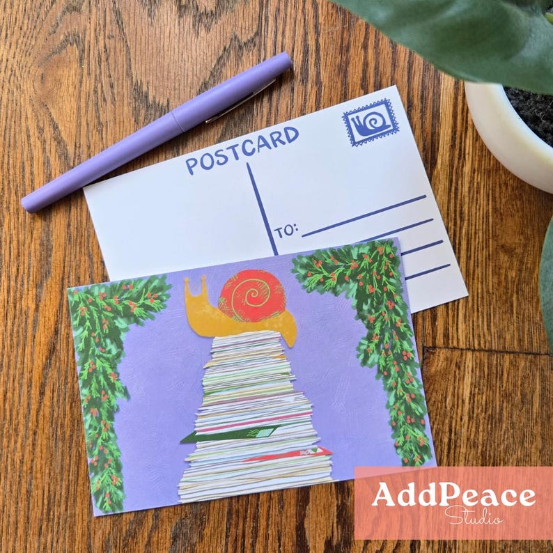 May include: A postcard featuring a snail illustration on a pile of books, surrounded by green foliage. A purple pen and a blank postcard with "POSTCARD" printed on it are also visible. The postcard has a blue stamp and lines for the recipient's address.