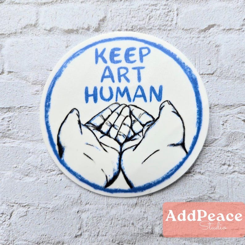 May include: A round, white ceramic art piece with a blue border and the text "KEEP ART HUMAN" in blue. The design features a pair of cupped hands cradling a stylized image of more hands. The bottom right corner has the text "AddPeace Studio".