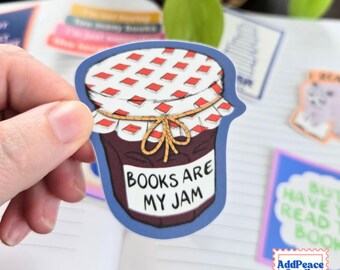 May include: A die-cut sticker shaped like a glass jar of dark purple jam. The lid is covered with a white cloth featuring a red diamond pattern, tied with a yellow string. A white label on the front reads, "BOOKS ARE MY JAM" in black text.