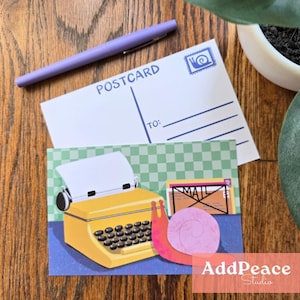 May include: A postcard and a greeting card featuring a yellow typewriter illustration, a pink snail, and the word "MAIL". A purple pen rests on the postcard. The background is a wooden surface.