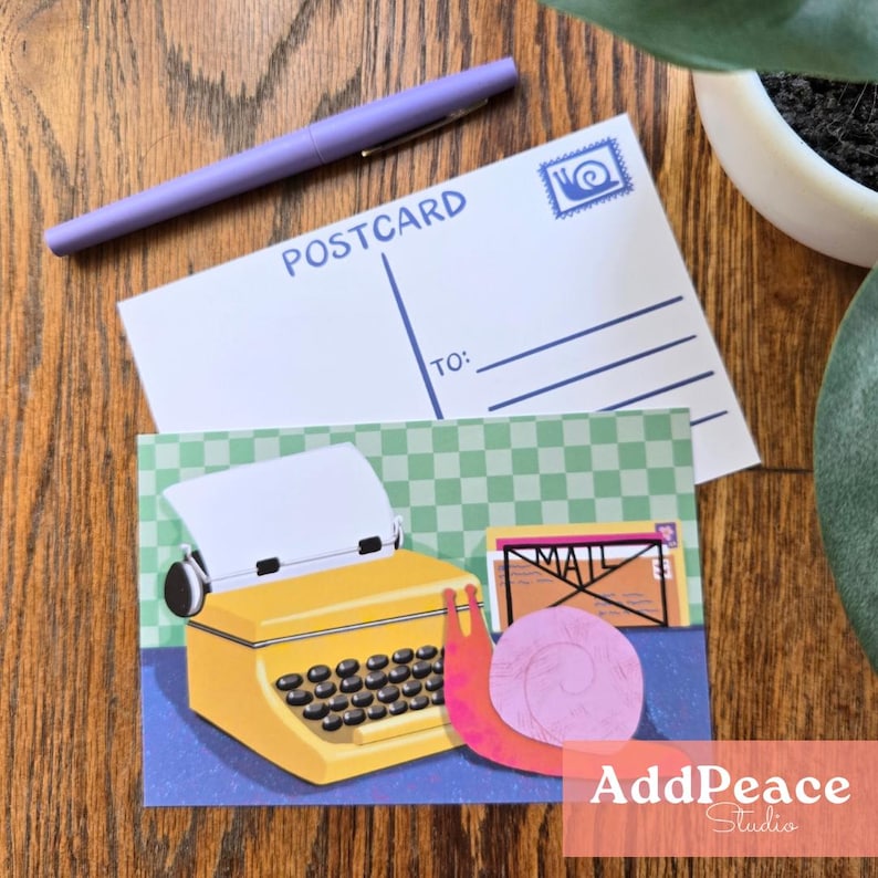 May include: A postcard and a greeting card featuring a yellow typewriter illustration, a pink snail, and the word "MAIL". A purple pen rests on the postcard. The background is a wooden surface.