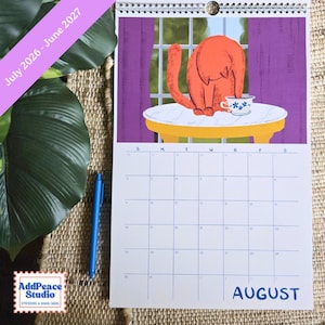 May include: A spiral-bound calendar featuring an illustration of an orange cat sitting at a table with a teacup. The calendar shows the month of August and the dates. Text on the calendar reads "July 2026 - June 2027". The calendar is on a woven surface.