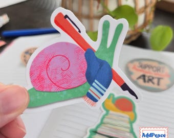 May include: A die-cut sticker featuring a green snail with a pink textured shell, wearing a blue scarf with white and red stripes, and carrying a long orange pen across its back. The sticker has a white border and a matte finish.