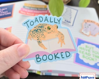 Op de afbeelding: Een gestanste sticker met een beige en groene pad die op een open boek met roze pagina's rust. De tekst TOADALLY BOOKED is in donkergroenblauwe letters boven en onder de pad gedrukt. De sticker heeft een lichtblauwe rand en een matte afwerking.