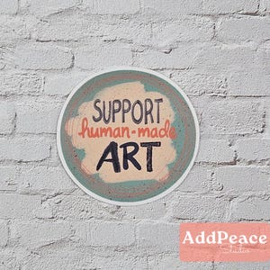 May include: A round sticker with a teal and peach background. The sticker has the text "SUPPORT human-made ART" in black and orange lettering.