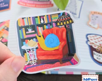 May include: A die-cut sticker featuring a yellow snail with a blue shell resting on a bright red armchair. The scene includes a white side table with a small paper, a dark bookshelf filled with colorful books, and a stained glass floor lamp.