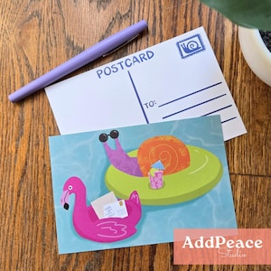 May include: Two postcards with quirky illustrations and a purple pen on a wooden surface. One postcard shows a flamingo with a letter, the other a snail with sunglasses. The word "POSTCARD" is printed on the blank postcard.
