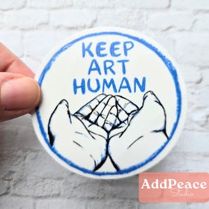 May include: A round, white sticker with a blue border and the words "KEEP ART HUMAN" in blue. The sticker features a drawing of two hands cupping a detailed drawing of a pair of hands.