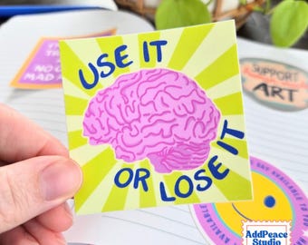 May include: A square sticker featuring a pink brain illustration centered on a lime green sunburst background. The text USE IT OR LOSE IT is printed in bold blue capital letters, curving around the top and bottom of the brain graphic.