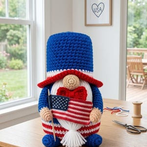 Peut inclure: Un gnome crocheté aux couleurs du drapeau américain, rouge, blanc et bleu, tenant un petit drapeau américain. Le gnome porte un chapeau bleu, une barbe blanche et un nœud papillon rouge. Il est posé sur une table en bois.