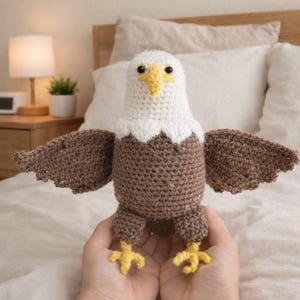 Peut inclure: Un aigle chauve fait à la main en crochet, les ailes déployées. L'aigle a une tête blanche, un bec jaune et un corps et des ailes bruns. Les serres de l'aigle sont jaunes. L'artisanat est présenté sur un fond doux et neutre.