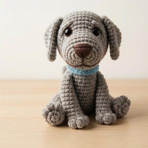 Peut inclure: Une figurine de chien crochetée grise avec de grands yeux marron foncé et un nez marron foncé. Le chien a des oreilles tombantes et porte un collier tressé bleu clair autour du cou. La texture du fil est visible sur toute la figurine.