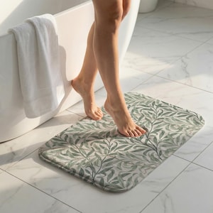 May include: A rectangular bath mat with rounded corners featuring a green and white botanical leaf pattern. The mat is placed on a tiled floor near a white bathtub. A white towel hangs on the side of the tub.