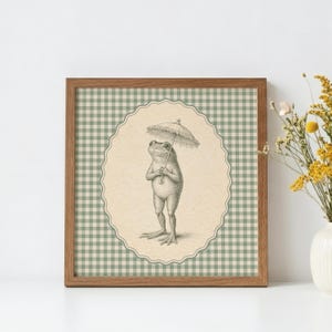 May include: A framed print featuring a detailed illustration of a frog holding an umbrella. The frog is standing upright, and the artwork is set against a cream-colored oval background with a green and white checkered pattern.