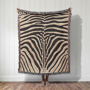 May include: A rectangular woven throw blanket featuring a repeating zebra stripe pattern in black and tan. The blanket has a textured appearance with decorative fringe detailing along all four edges, creating a distinct border around the animal print.