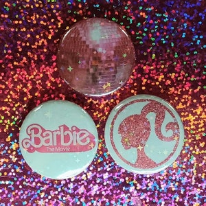 May include: Three round, light blue, pinback buttons with a pink glitter design. The first button has a disco ball design. The second button has the text "Barbie The Movie" in pink glitter. The third button has a pink glitter silhouette of a woman with a ponytail.
