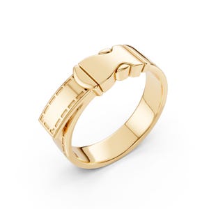 Puede incluir: Un anillo dorado con diseño de hebilla. El anillo presenta una placa rectangular con detalles cosidos y un cierre. El anillo está hecho de un metal brillante y reflectante.