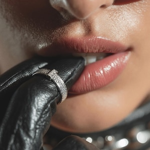 May include: Close-up of a silver ring with a square center stone and a band covered in small stones. The ring is on a black leather glove. The person is wearing sunglasses and has glossy lips.