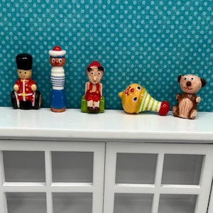 May include: A collection of miniature wooden toys, including a soldier, a sailor, a figure in red, a yellow and green noisemaker, and a small bear. The toys are displayed on a white cabinet against a teal background with white polka dots.