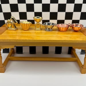 May include: Miniature kitchen tools on a wooden table. The set includes a gold mixer, a fluted bowl, a scale, a silver bowl, and two copper bowls. The background is a black and white checkered pattern.