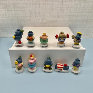 May include: A collection of ten small, ceramic hippo figurines, each with a unique design and pose. The hippos are painted in various colors and feature different accessories, such as a guitar, dumbbells, and a chef's hat. The figurines are displayed on a white rectangular base.