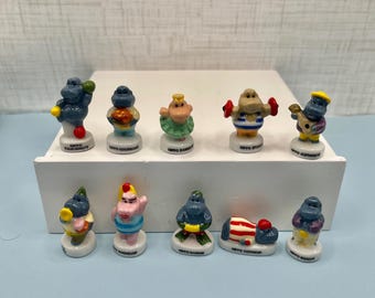 May include: A collection of ten small, ceramic hippo figurines, each with a unique design and pose. The hippos are painted in various colors and feature different accessories, such as a guitar, dumbbells, and a chef's hat. The figurines are displayed on a white rectangular base.