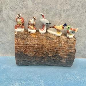 May include: A collection of ceramic bird figurines, including an owl, eagle, stork, pheasant, and duck, displayed on a rustic wooden log. Each bird is painted with detailed colors and rests on a base with French text.