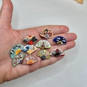 May include: A collection of eleven small, semi-circular fan-shaped ornaments featuring diverse artistic designs, including depictions of waves, pagodas, floral patterns, and figures, rendered in various colors such as blue, gold, green, and red.