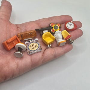 May include: A collection of miniature furniture and decor items, including a small orange sofa, a wooden cabinet, a yellow chair, a white bust, a red clock, and a lamp. The items are held in a hand.