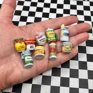 May include: A collection of nine miniature ceramic jars and containers featuring various colorful labels and painted details. The items include small pots labeled with text such as Fruits a la Liqueur, Bonbons, Foie Gras, and Legumes, showcasing diverse shapes.
