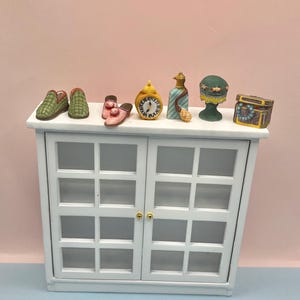 May include: A white cabinet with glass-paned doors displays miniature decorative items. These include green and pink slippers, a yellow clock, a blue bottle, a green hat, and a small treasure chest. The items are arranged on top of the cabinet.