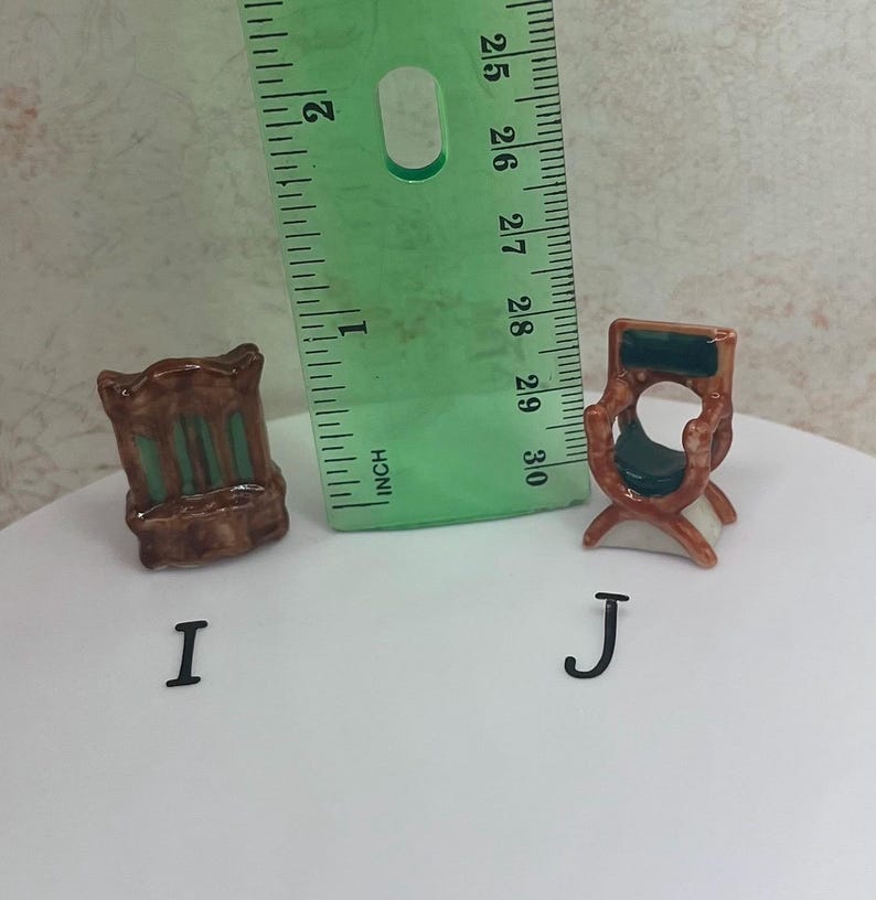May include: Two miniature ceramic chairs, one with a brown frame and green accents, the other with a brown frame and a green seat. A green ruler is placed next to the chairs. The chairs are labeled with the letters "I" and "J".