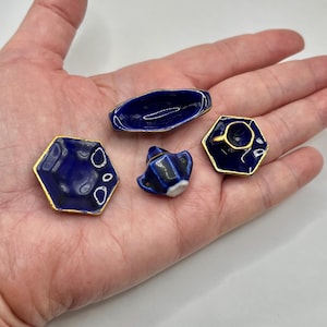 May include: Miniature blue ceramic tea set with gold trim, including a hexagonal plate, a small teapot, a long dish, and a teacup with a saucer. The set is displayed on a hand, showcasing the small size of the pieces.