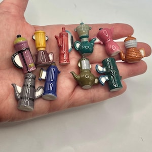 May include: A collection of eleven miniature ceramic coffee pots and teapots in various colors including pink, blue, green, yellow, brown, and grey. Each small vessel features a distinct handle, spout, and lid design, showcasing a glossy finish.