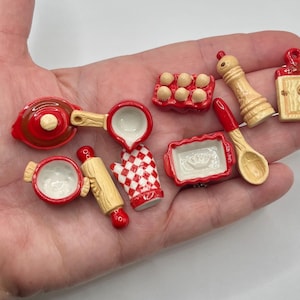 May include: Miniature kitchen accessories in red, white, and tan. The set includes a saucepan, measuring cup, rolling pin, oven mitt, baking dish, egg holder, salt shaker, spoon, and cutting board. These small items are likely for a dollhouse or craft project.